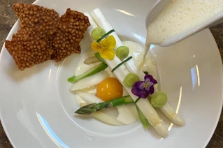 Gourmet aspargesret med pocheret æg, knasende brødkiks, spiselige blomster og sauce bliver hældt på.