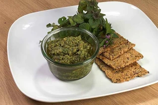 Hjemmelavet pesto i glas med krydderurter og knækbrød på hvid tallerken.