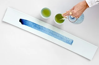 Person hælder grøn te fra tekande i traditionelle japanske te kopper, minimalistisk borddækning.