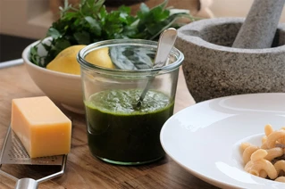 Friske ingredienser klar til madlavning: grøn pesto i glas, parmesanost, citron, persille, hvidløg og morter på køkkenbord.