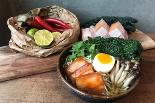 Skål med ramen nudler, blødkogt æg, svampe og grøntsager, med friske krydderier og skiver af lime ved siden på en træskærebræt, ideel til opskriftsider og madblogs