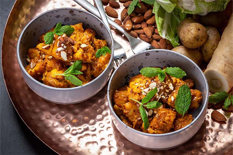 To skåle med krydret græskarret toppet med hakkede mandler og frisk mynte, præsenteret på et kobberfad med hele mandler og grøntsager i baggrunden, der fremhæver sund vegetarisk madlavning.