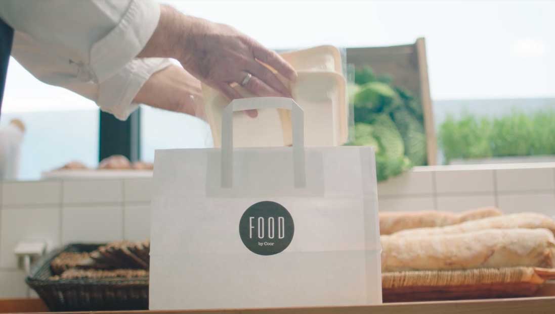 Person overdrager hvid take-away pose mærket 'FOOD by Coor' over skranke i moderne køkken