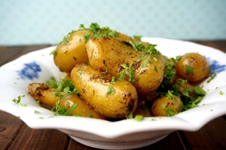 Friske ovnbagte kartofler drysset med finthakket persille serveret i en hvid skål med blåt mønster, perfekt som tilbehør til middagsretter.