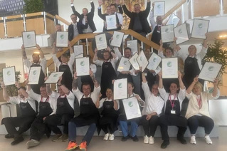 Gruppe af glade mennesker der fejrer med diplommer og iført kokkeforklæder i et lyst kontormiljø.